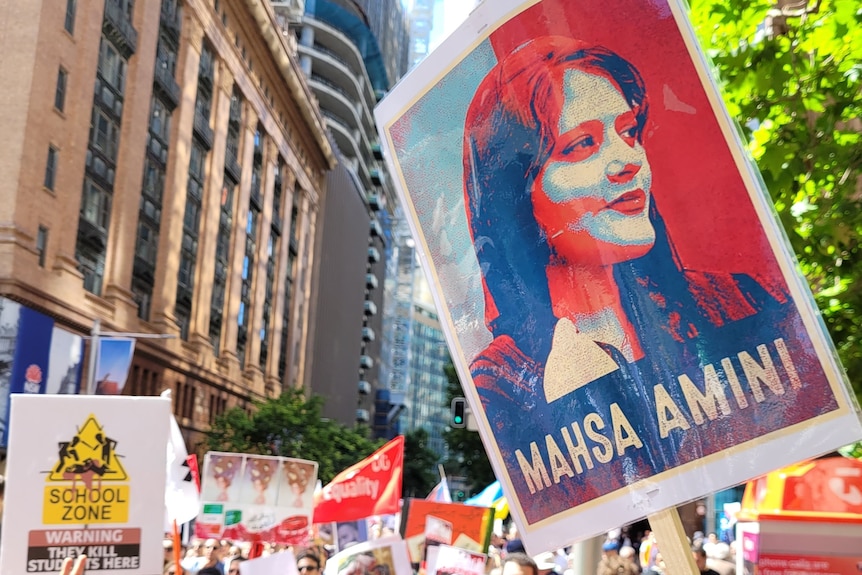 A blue, red and white sign of a woman in a hijab, with the words Mahsa Amini, as protesters gather in the background