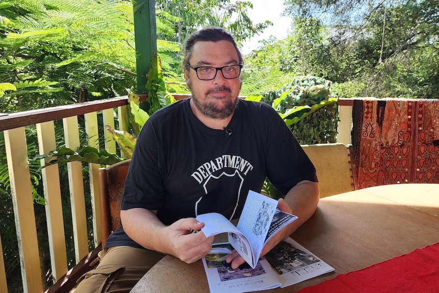 A man holds a small printed magazine while sitting on an outdoor verandah.