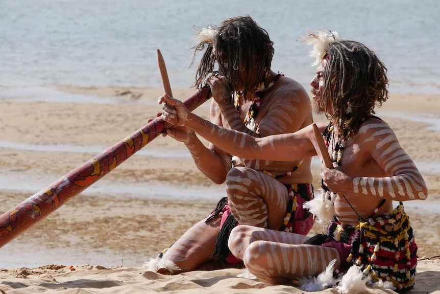 Two Aboriginal men play traditional indigenous instruments on a beach while wearing traditional paint and wear.