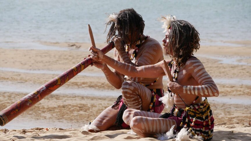 Two Aboriginal men play traditional indigenous instruments on a beach while wearing traditional paint and wear.