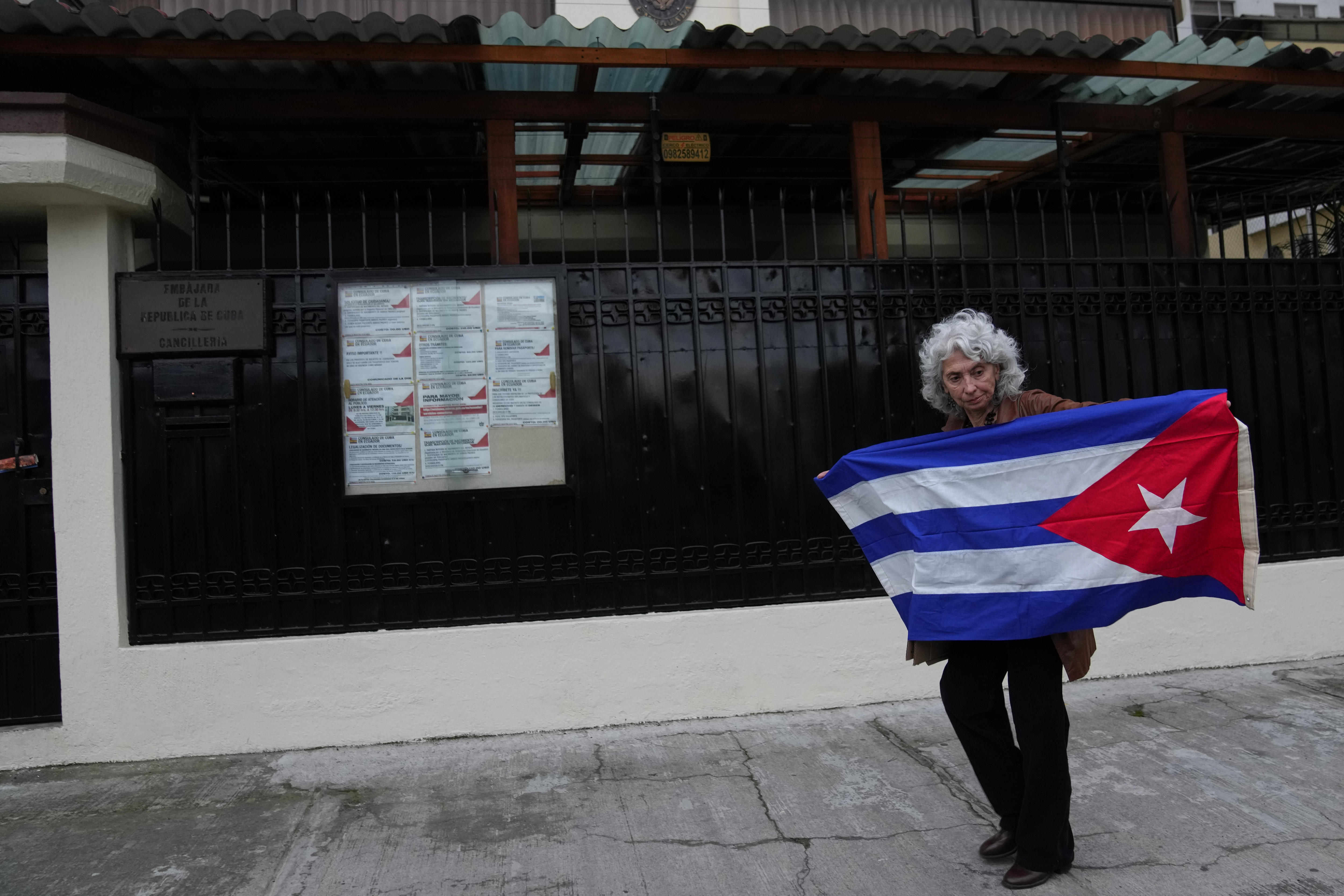 Una mujer de pelo gris sostiene una bandera cubana en un camino junto a una gran valla de metal negro.