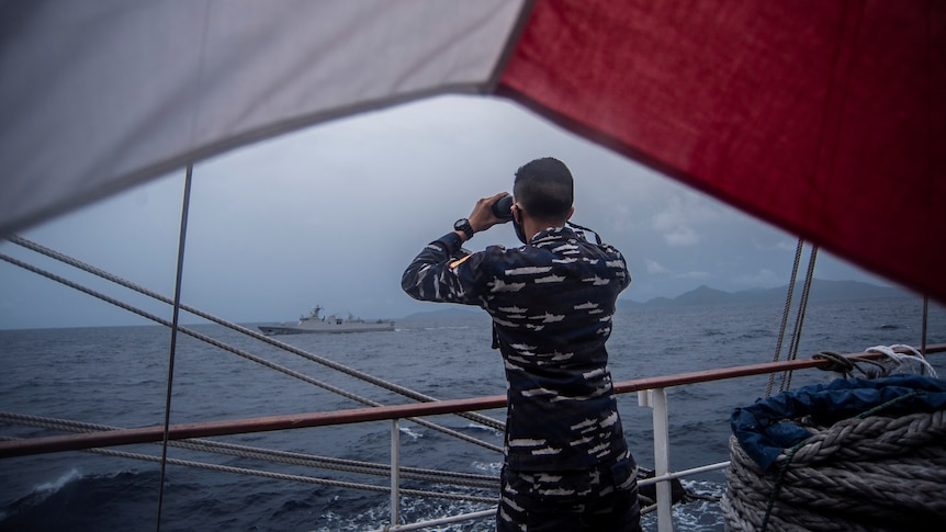 A Navy officer in camo fatigues uses binoculars to look from the side of his ship towards another boat across choppy grey water