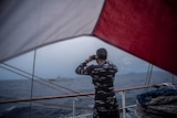 A Navy officer in camo fatigues uses binoculars to look from the side of his ship towards another boat across choppy grey water