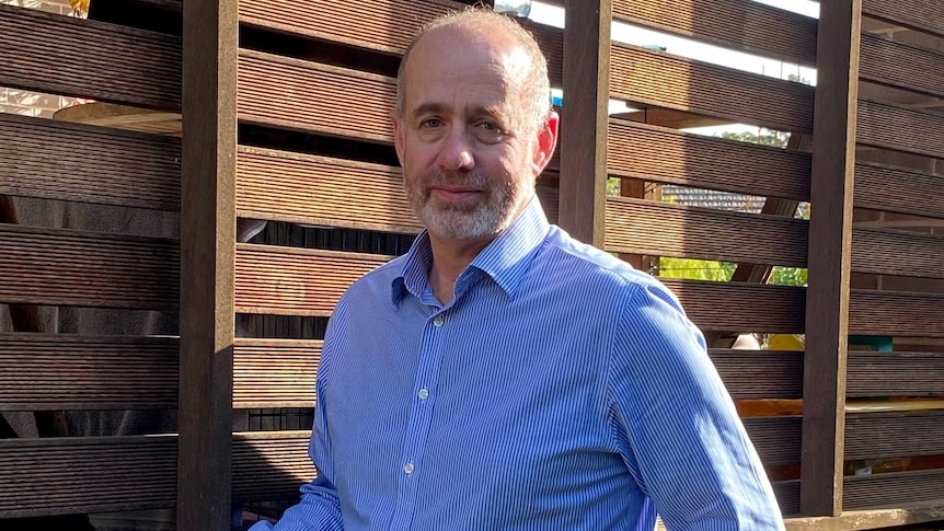 bearded man wearing a striped collared shirt standing in front of tall timber fence