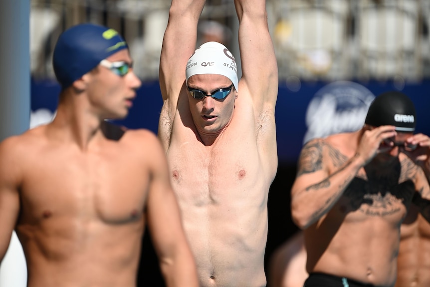 Man in swimming cap stretches his arms in the air, with two other men in caps either side of him.