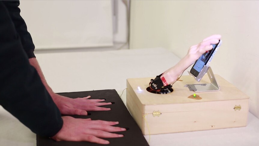 A man holds his hands on a sensor plate while a robot hand operates a smartphone screen.