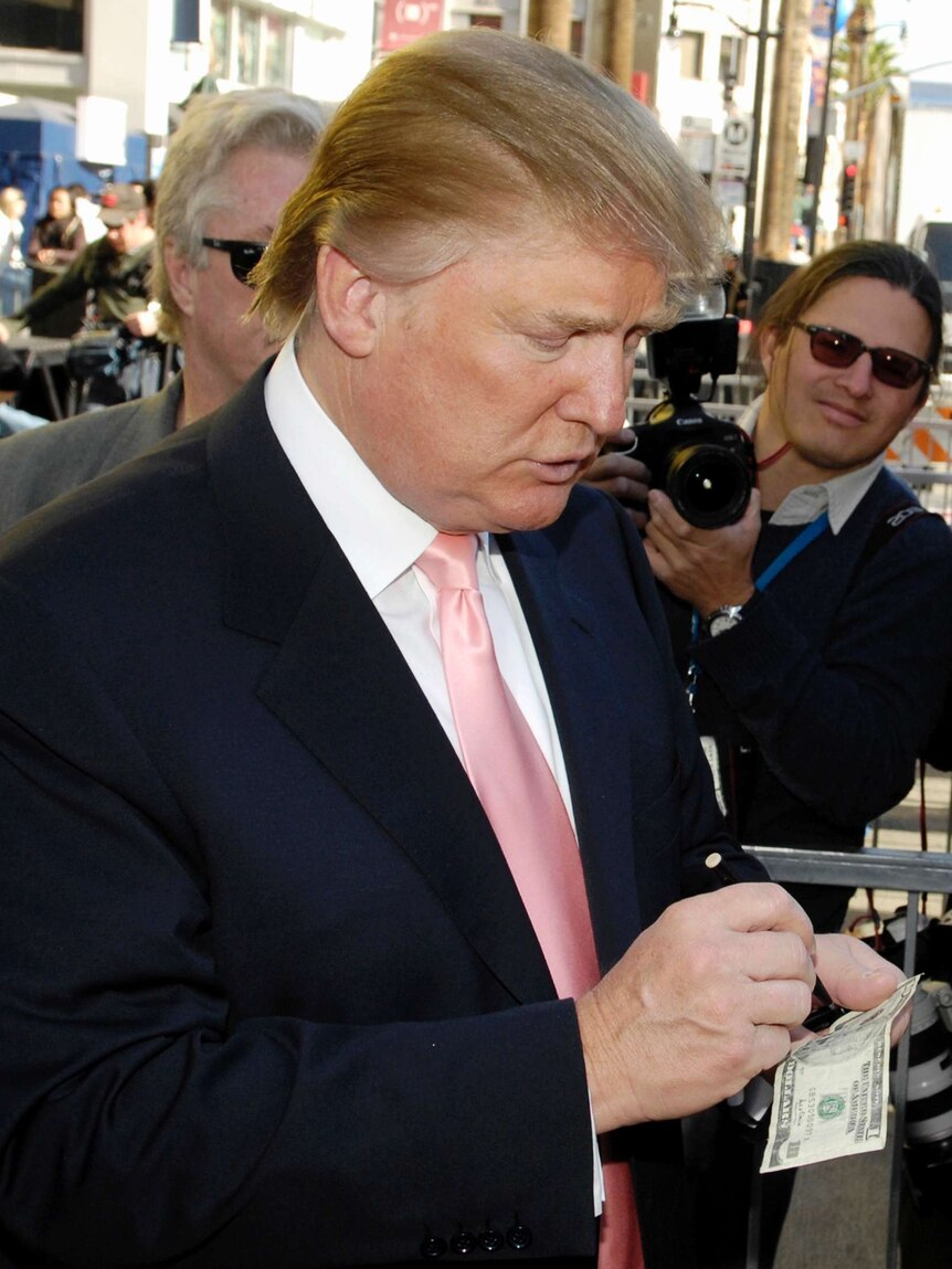 Donald Trump looks down at an American dollar bill and signs it for a fan.