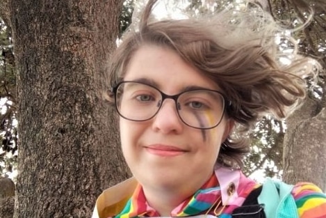 A person with flowy short brown hair wearing black glasses and colourful rainbow top and facepaint 