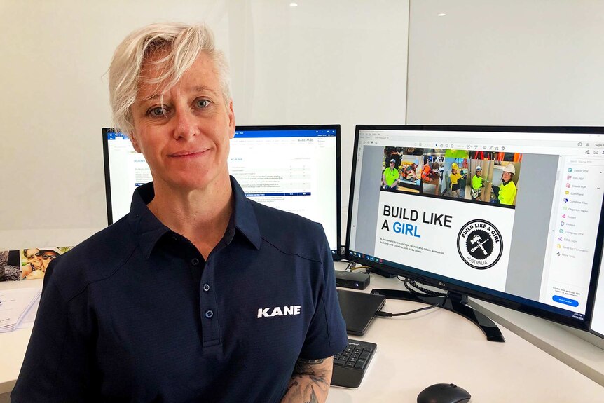 A woman with very short blonde hair wearing a navy polo shirt with the word Kane written on it stands in front of a computer.