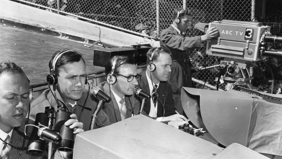 Four men with microphones and headphone sit outside next to a man with a very large television camera.