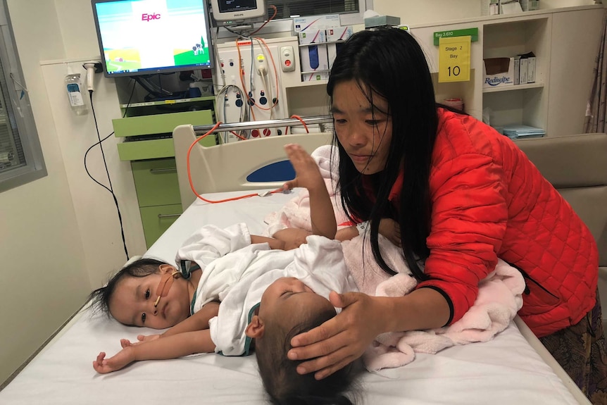 Two toddlers lie in a hospital bed, comforted by their mother