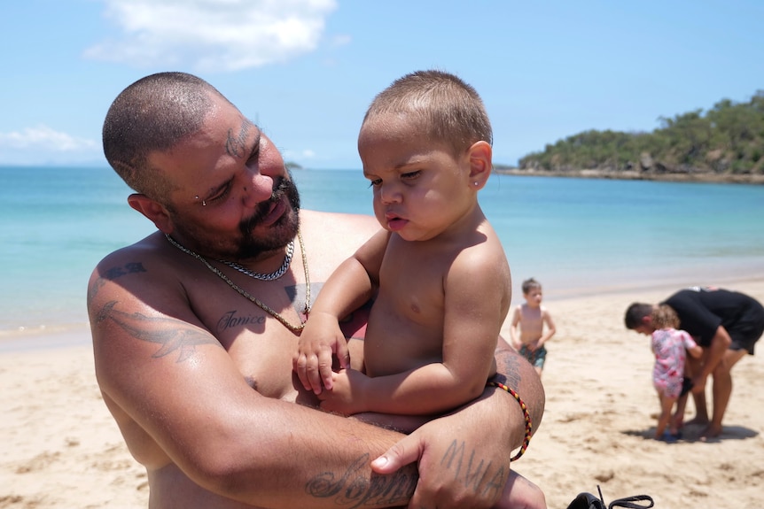 An Indigenous man holding an Indigenous baby on an Island, smiling at him. 