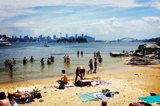A group of people gather on the sand and in the water at a beach, with Sydney Harbour Bridge visible on the horizon.