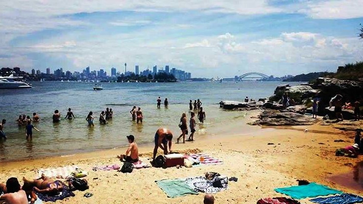 A group of people gather on the sand and in the water at a beach, with Sydney Harbour Bridge visible on the horizon.
