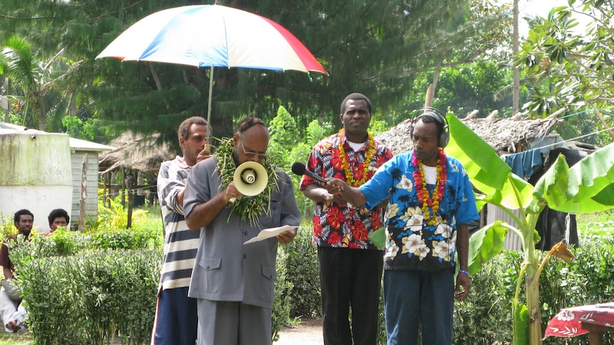 Vanuatu politician speaks through a megaphone with media holding microphone and umbrella