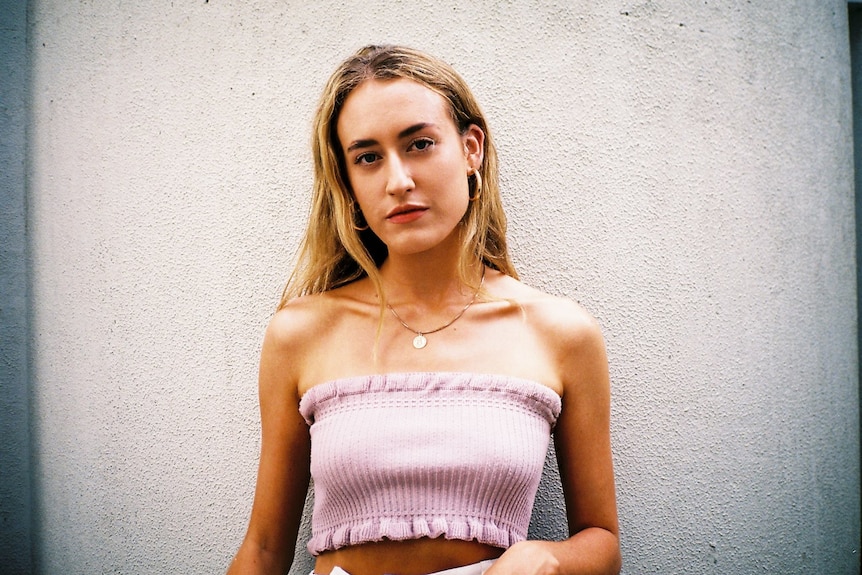 A woman with blonde hair stands against a white wall wearing a light pink top. 