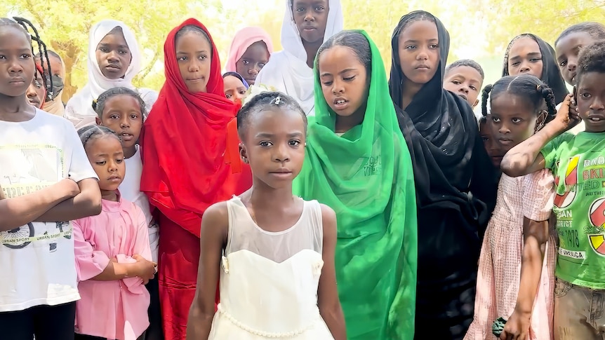 A serious Sudanese girl stands in the centre of 17 other kids  who have come to listen to her poetry.