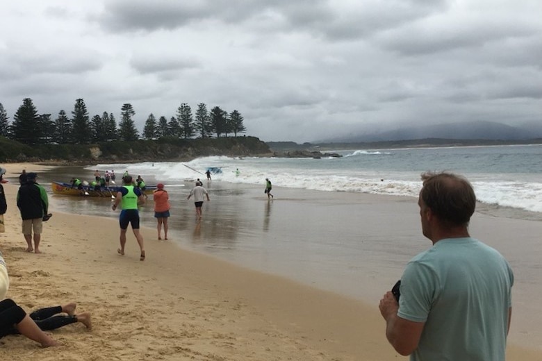 An overcast day at the beach. Spectators stand on sand, people in high-vis rush towards upturned boat in shallow water