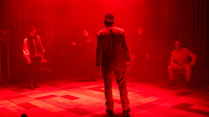 Bathed in foreboding red light, a man wearing a crown faces a group of figures in shadow. 
