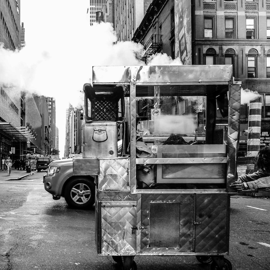 A black and white photo shows a street vendor pushing a trolley through a New York intersection, with steam rising behind him.