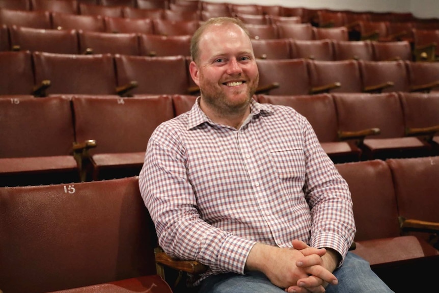 A man in a checked shirt smiles at the camera seated in a row of red cinema chairs