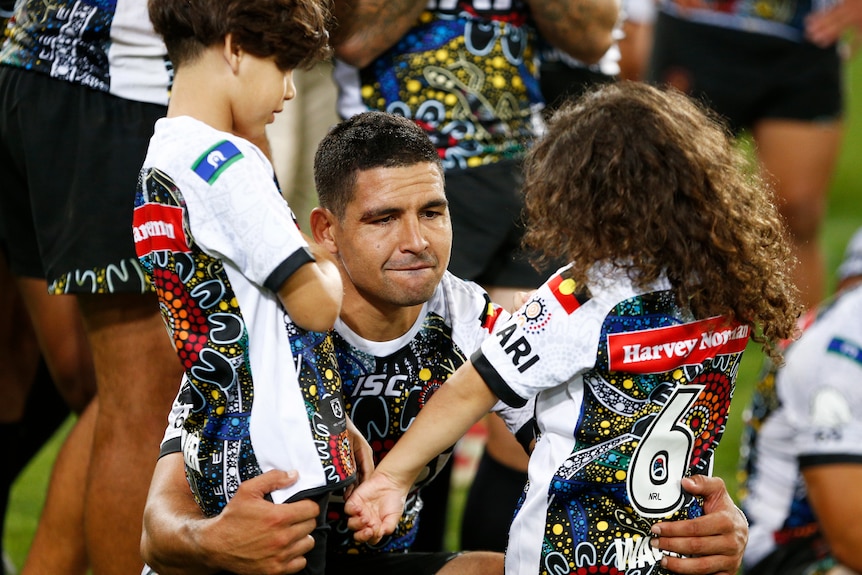 Cody Walker crouches down hugging his two children, standing up, with one in each arm