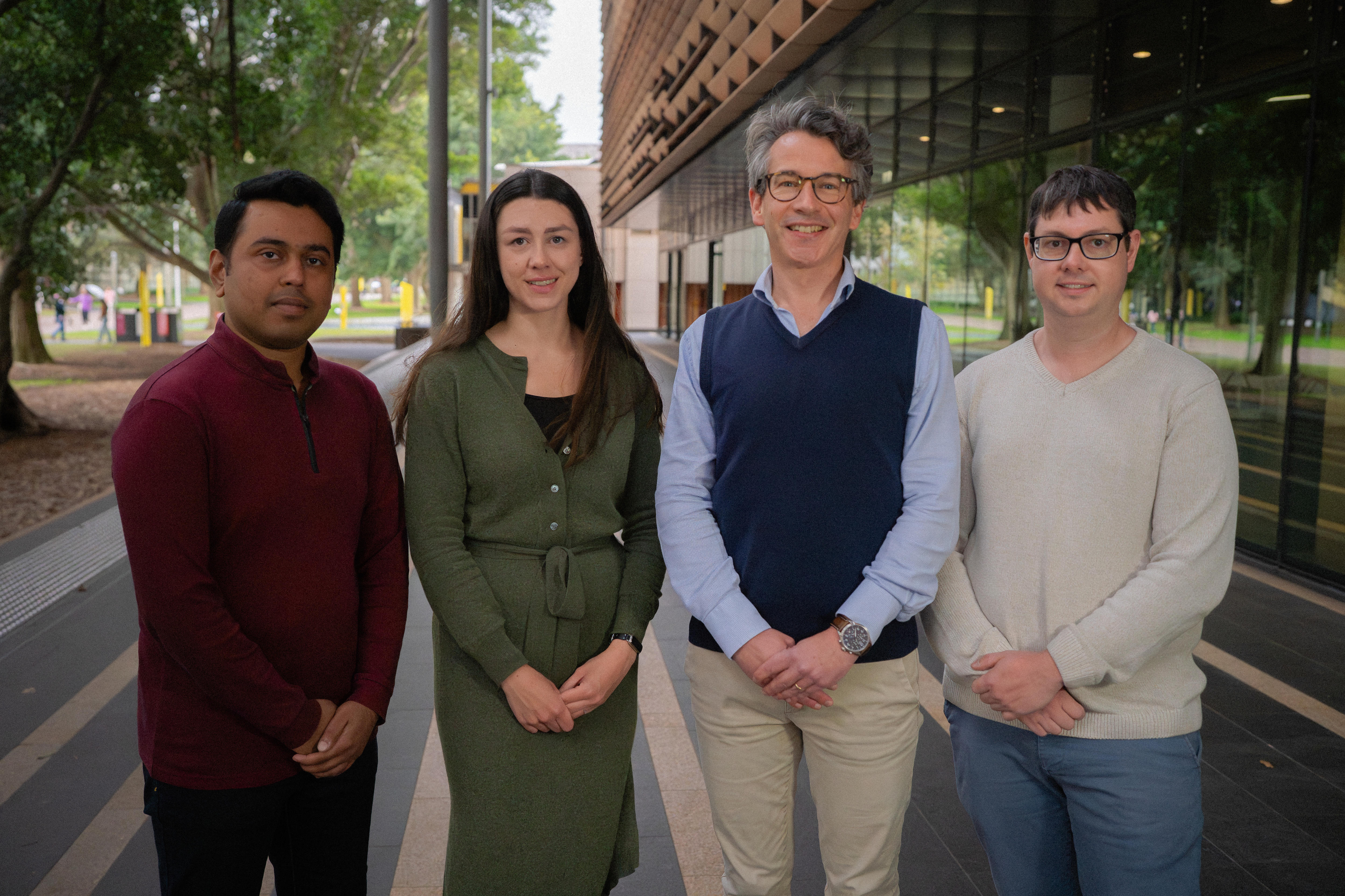 A team of four researchers - one woman and three men - smile and hold their own hands as they stand together outside a building.