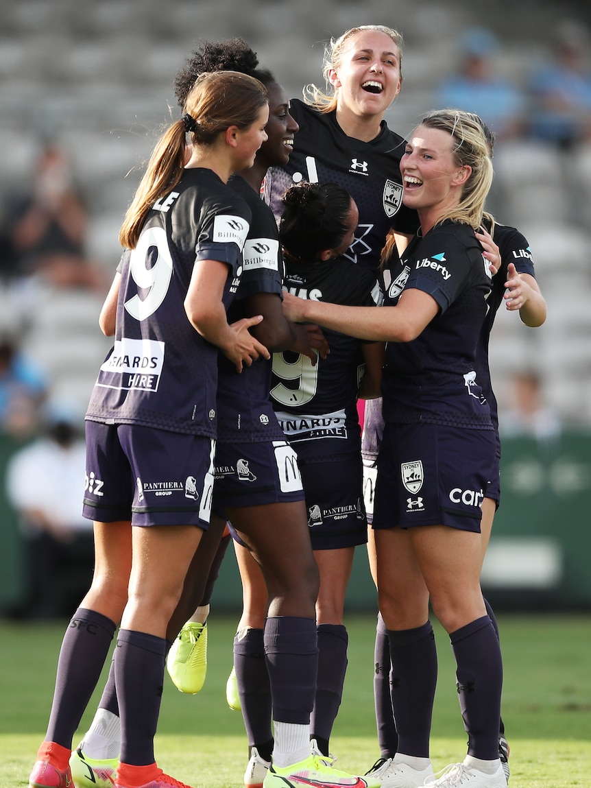 Women soccer players wearing dark blue hug each other after scoring a goal