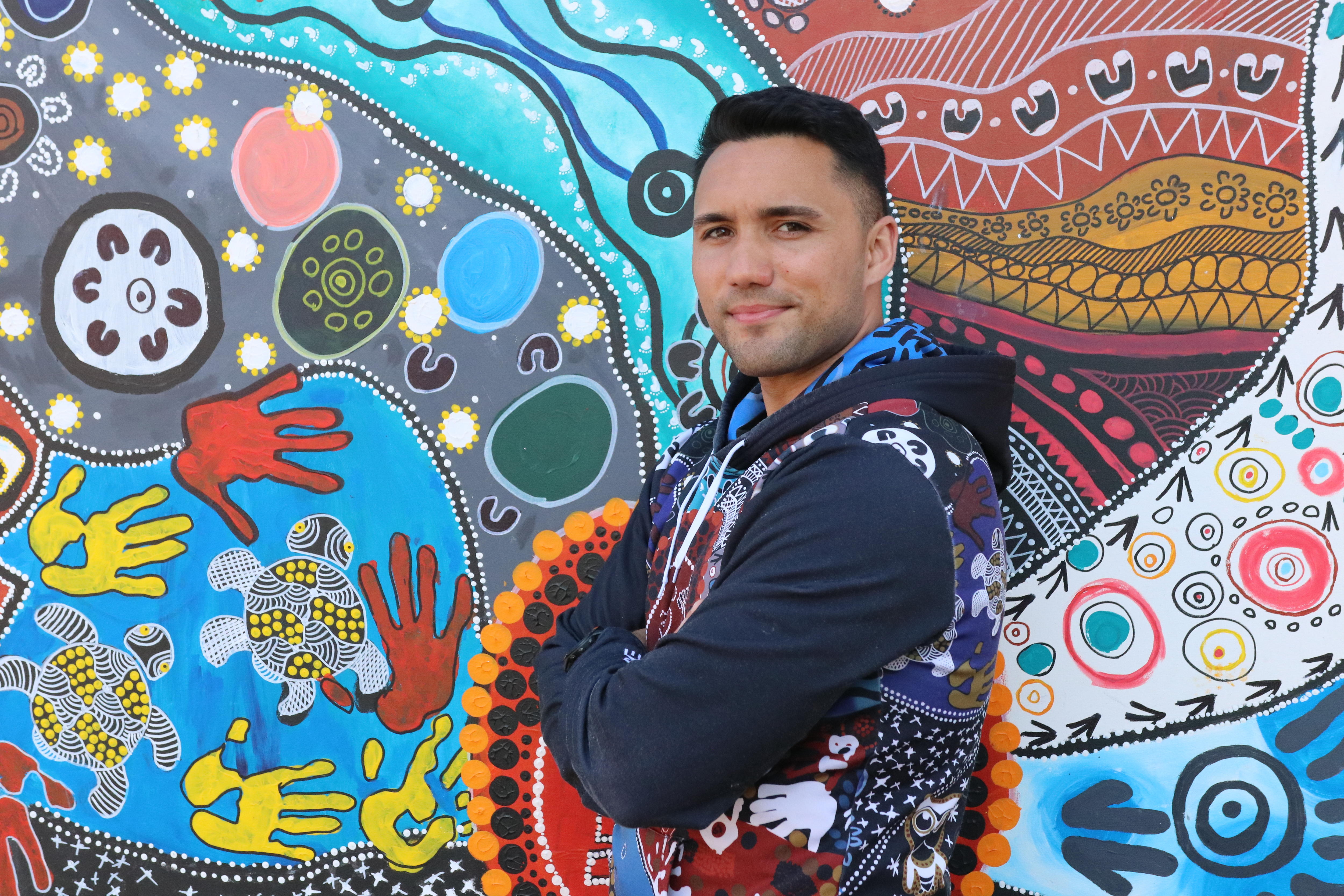Isaiah Dawe is standing with his arms crossed, smiling. He is wearing a hoodie, standing in front of a bright Indigenous mural