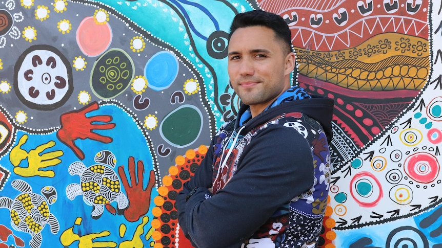Isaiah Dawe is standing with his arms crossed, smiling. He is wearing a hoodie, standing in front of a bright Indigenous mural