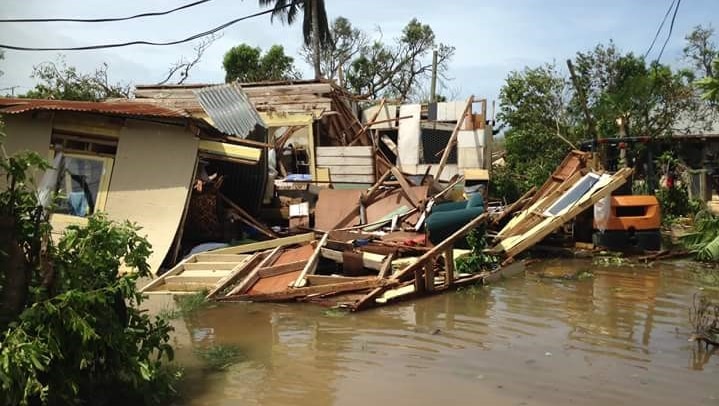 A low-set home sits destroyed in murky flood waters, a windswept palm tree is in the background