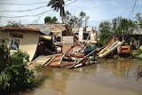 A low-set home sits destroyed in murky flood waters, a windswept palm tree is in the background
