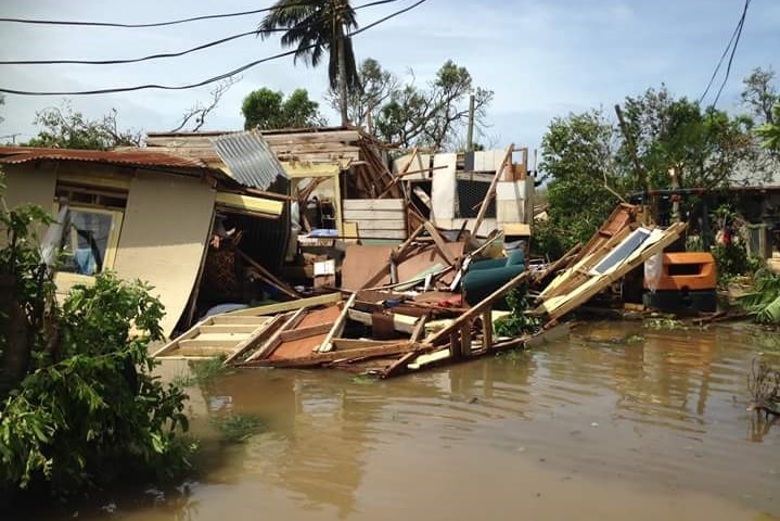 A low-set home sits destroyed in murky flood waters, a windswept palm tree is in the background