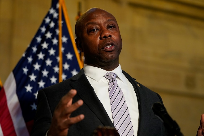 A middle-aged, bald black man in a suit speaks in front of an American flag.