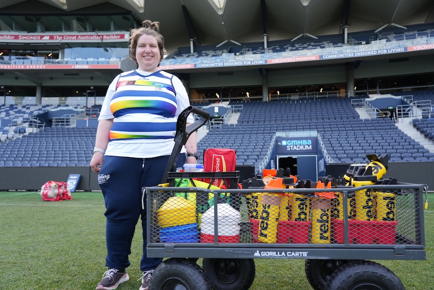 Belinda Cini wearing a Geelong Cats Pride Jersey, standing on an AFL field next to a water cart.
