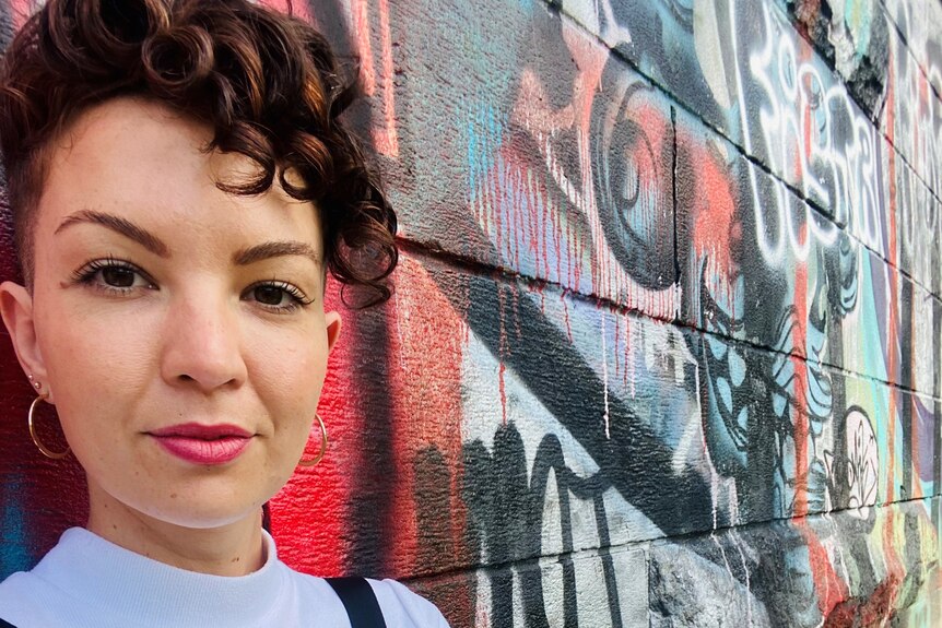 Sarah McCarthy smiles while standing with her back again a wall.