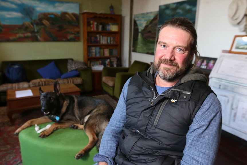Adam Stow sits inside wearing a sleeveless puffer jacket and blue shirt with his dog sitting nearby.