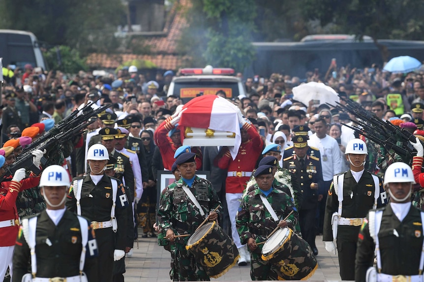A procession of soldiers with drums walk before a coffin, followed by a crowd of people in military uniforms.