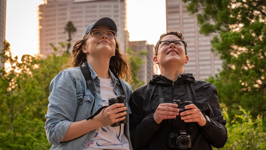 A photo of Amy Ranck and Stephanie Chambers looking up a some birds, holding their binoculars.