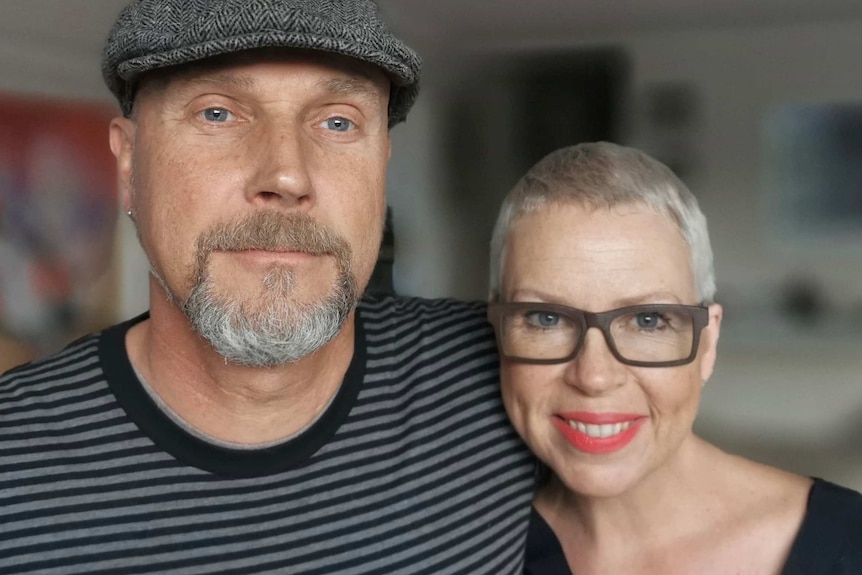 A man with a flat cap and short grey beard has his arm around a woman with short grey hair and thick-rimmed glasses.