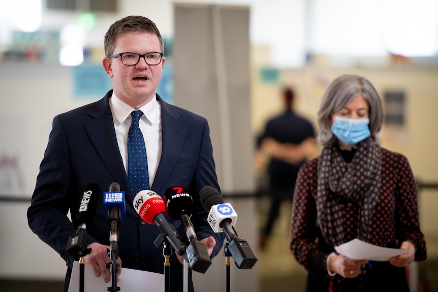 A man wearing glasses speaking into microphones with a woman wearing a mask in the background 