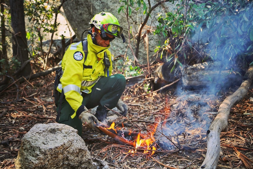 A man in high-vis lights a fire in a forest.