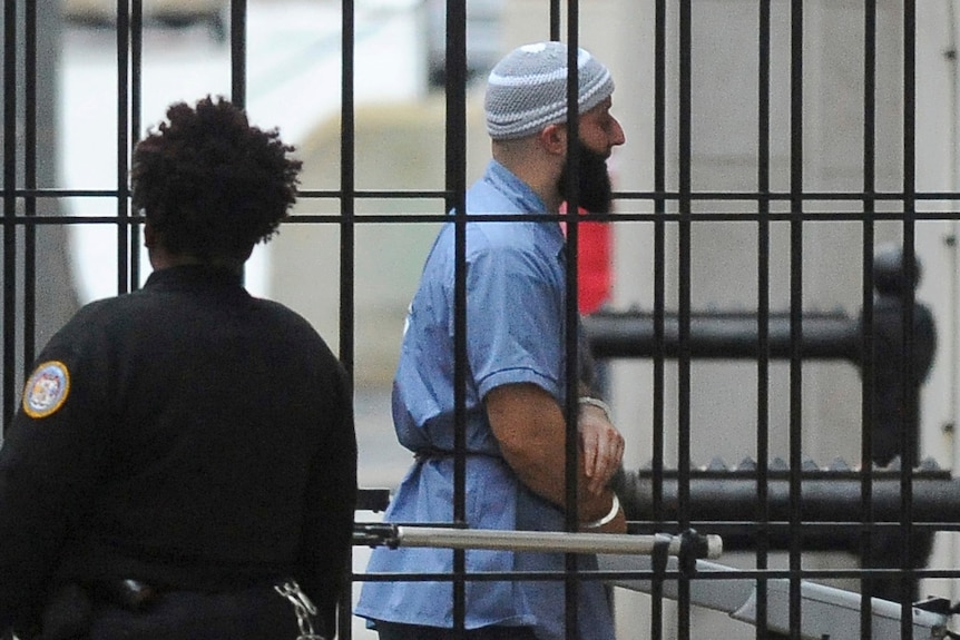 adnan, in prison uniform and with his head covered, walks behind bars, handcuffed