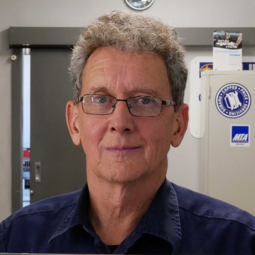 a man with glasses looking at the camera with an office in the background