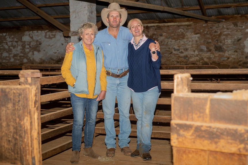 Man and two women stand arm in arm in a rustic shearing shed. 