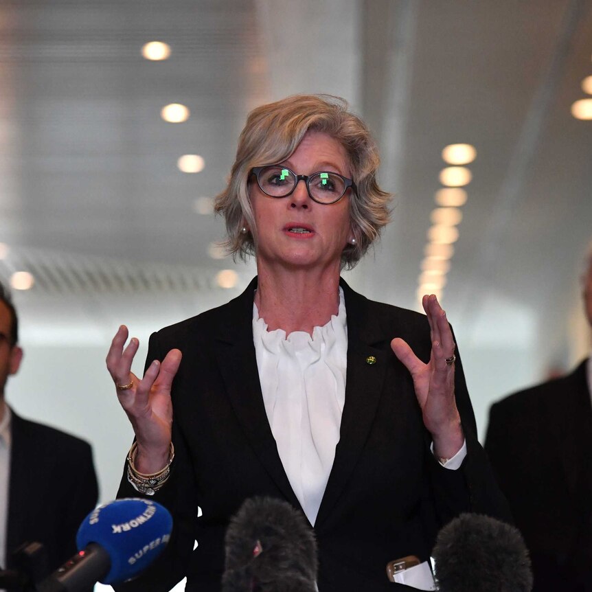 A woman in glasses and a black suit jacket gives a press conference inside with two men standing behind her.