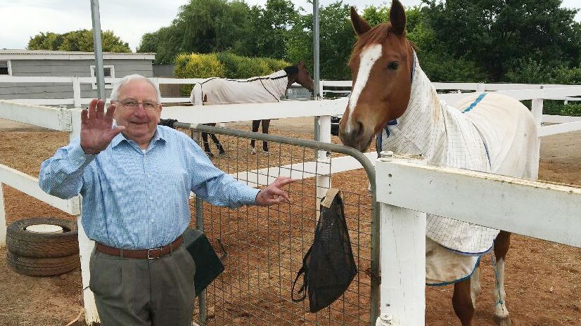 Mick Robins standing by a white horse enclosure holding his hand out with five fingers