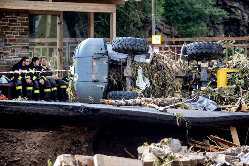 Firefighters sit on a bench outside a house near a truck covered in detritus on its side.