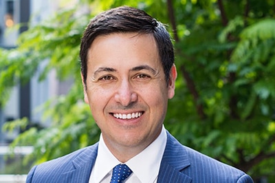 A smiling Keith John in a blue suit and tie stands in front of a tree.