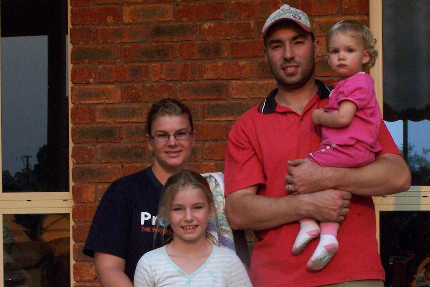 Zach holding toddler daughter and Brooke with arms around primary-school aged daughter standing outside their house.